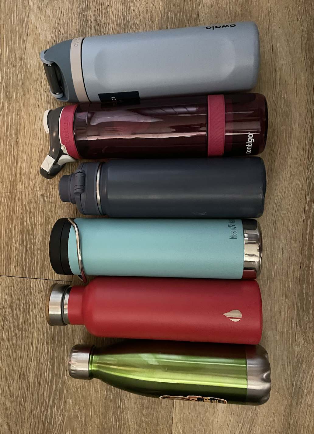 Top to bottom: A light blue water bottle with a dark blue lid, a transparent red water bottle, a dark grey metal water bottle, a light blue water bottle with a black lid, a red metal water bottle with a wooden lid, a light green water bottle